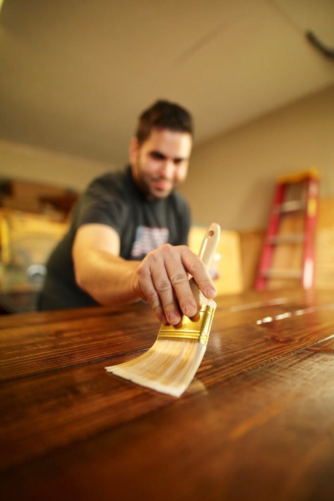 A man carefully applies varnish to a wooden surface indoors, using a paintbrush for a smooth finish.