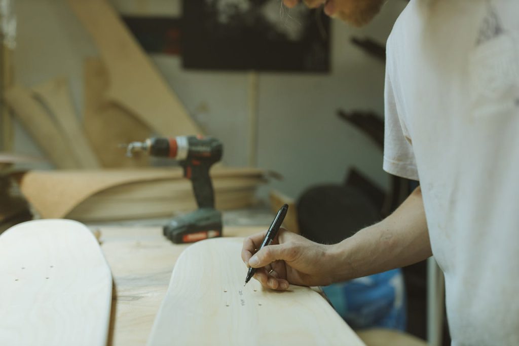 A craftsman in a workshop marks a skateboard deck with precision tools, surrounded by wood and drill.